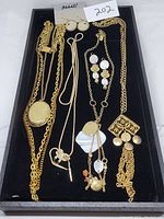 Goldtone necklaces and earrings arranged on a black velvet display tray, featuring a locket necklace, lariat necklace signed Sarah, matching earring set, and decorative necklace with round pendants.