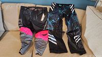 Two pairs of Fly motorcycle pants, one black with pink and gray accents, the other black with blue and gray graphic patterns and white stripes.