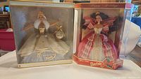 Two vintage holiday Barbie dolls in original unopened packaging standing side by side. Celebration Barbie in gold and white gown, Happy Holidays Barbie in red and white gown.