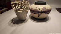 View showing both pottery pieces side by side on a flat surface under soft light; highlights ivory cutout vase and round raku vessel with plum bands.