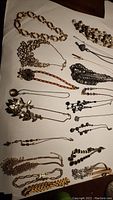 Assorted necklaces laid out on a white surface, showing a variety of chain styles, lengths, beads, and pendants including floral, heart, leaf, and abstract designs.