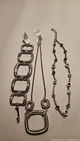 Photo shows three pieces of Brighton jewelry laid flat against a beige background. Bracelet with rectangular textured links, necklace with large square pendant and two small squares on chain, and a crystal-accented choker are visible.