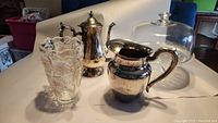 Silver plated water pitcher, coffee pot, vintage glass vase, and glass cake plate with dome shown together on a table.