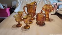 Full view of all 7 pieces of amber carnival glass including pitcher, glasses, candy jar, and fluted vase, all with raised grape design and iridescent finish.