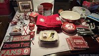 Alabama-themed lot of various items laid out on table including towels, plates, cups, fan sign, and serving bucket.