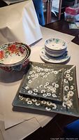 Photo showing 2 square serving trays with olive green and white cherry blossom design, 4 floral mixing bowls with red and pink flowers, and 4 place settings with blue floral small plates and bowls.