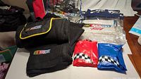 Photo showing two black Nascar branded blankets, two clear Nascar tote bags, one clear Nascar backpack, and two inflatable seat covers in red and blue with checkered flag patterns.