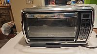Full view of Oster toaster oven showing stainless steel body, glass door, and handle, positioned on a white surface.