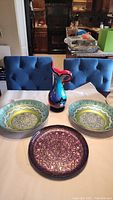 Two blue, silver, and green Moroccan style glass serving bowls, purple and gray glass serving tray, and multicolored Murano style glass pitcher arranged on a table.