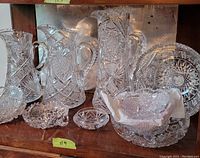 Photo of the assortment of large cut glass pitchers and bowls displayed on a wooden shelf. Includes detailed views of multiple pitchered and bowls with intricate cut glass patterns.