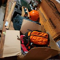 View of a cardboard box containing an orange backpack and a dark green duffel bag with a bright orange helmet behind it.