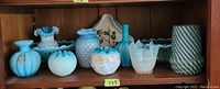 Shelf with nine blue collectible glass items including assorted vases and bowls displaying varied textures such as ruffled edges, bubble pattern, swirl design, and floral painting.