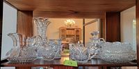 Photo showing seven assorted clear cut glass pitchers and trays arranged on a wooden shelf, reflecting light and displaying detailed cut patterns.