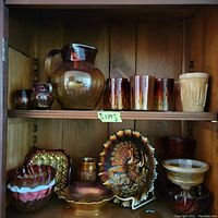 Wide view of upper and lower shelves displaying a variety of amber and carnival glass items including pitcher, glasses, decorative plate, and bowls.