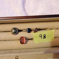 Front and angled view of four vintage rings with various stones including green, dark blue, raspberry and pinkish-orange on a beige ring holder.