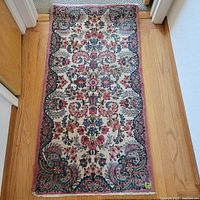 Photo of the handwoven scatter rug laying flat on wooden floor between two doorways. Shows the intricate floral and curved ornamental patterns in pink, blue, cream and black.