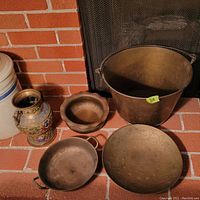 Photo showing all copper vessels arranged against brick background including bucket, two pans, scalloped bowl, decorative vase, and engraved plate