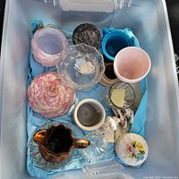 Top view of plastic container showing various ceramic, porcelain, and glass pieces including bowls, vases, dishes, and figurines arranged on blue tissue paper.