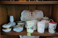 Porcelain and glassware lot including floral painted tray, teapot, stick pin holders, and tumblers arranged on wooden shelf.