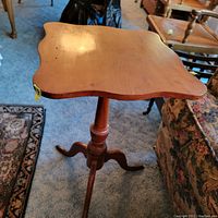 Front perspective of small wood side table showing curved edges of square top, turned pedestal, and three curved legs on carpeted floor.