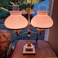 Twin lamp with lit ribbed milk glass shades on brass arms and marble base, placed on wooden table near window.