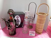 Photo showing all four items in the lot including glass decanter with stopper, metal golfer bottle holder, round metal wire bottle holder, and tall wicker basket holder