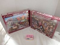Two boxed Christmas puzzles, Village Square and Christmas Kitchen, displayed side by side showing front of boxes