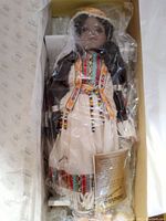 Porcelain doll in original packaging showing hand painted face and intricate traditional American Indian clothing with beads and detailed patterns.