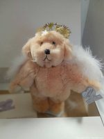 Front view of plush bear doll showing light tan fur, golden star halo, white feather wings, and embroidered facial features.