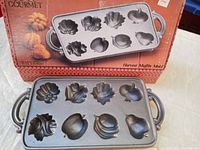 Top-down view of the cast iron muffin pan showcasing 10 harvest-shaped molds including leaves and fruit shapes, with handles on both sides and the product box in the background.
