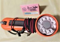 Side view of vintage orange lineman rotary telephone line tester with coiled black and red wire on handle and Rotary dial face with numbers and letters visible
