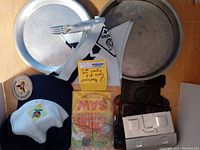 Top-down photo showing all items including 4 metal plates, fork, bandana with emblem, Boy Scout patch, camping stove parts, folded saw in packaging, and two hats.