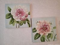 Two square canvas paintings with large pink flowers and green leaves hung on a wall, showing the full view of both pieces.