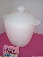 Front view of white hobnail milk glass ice bucket with lid and side handles on pink background.