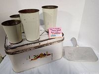 Photo showing a white vintage metal bread box with colorful painted fruit on front, four tall metal flower shop vases marked G. Savarese, and a clear plastic lid.