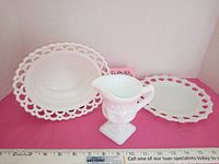 Lot of 3 vintage milk glass items shown together on a pink surface with measurement tape for scale. Includes creamer, lace edge bowl and lace edge plate.
