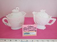 Two white milk glass creamers with lids, one swan-themed and one floral fruit basket-themed, positioned with a ruler for size reference.