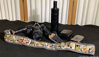 Full view of camera strap with multiple collectible enamel pins attached, along with Asahi Pentax MV1 film camera, Canon IXY digital camera, Nikon Coolpix case, and Celestron 9x50 finder scope arranged on flat black surface.