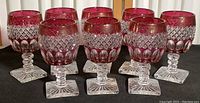 Overview photo of eight cranberry pressed glass goblets with clear stems and square bases arranged in two rows.