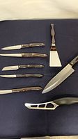 Eight items arranged on blue cloth: five wood-handled knives at left, offset spatula top right, chef’s knife and Santoku at bottom right.