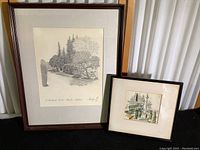 Two framed prints on black surface with white curtain background: larger etching on left, smaller watercolor on right