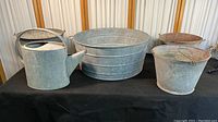 Full view of collection including galvanized watering can, large round tub, and three different sized buckets on black surface.