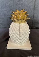 Front view of white and gold ceramic pineapple bookends placed side by side to form the shape of a whole pineapple.
