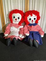 Both Raggedy Ann and Raggedy Andy dolls sitting side by side against a white paneled backdrop, showing full front view of the dolls.