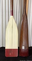 Side-by-side view of two large wooden oars showing different paddle shapes and colors, highlighting condition and design details.