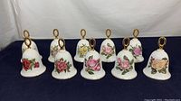 Full view of 10 vintage Danbury Mint bone china bells with American Rose floral designs arranged in a row on dark fabric background.