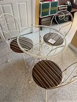 Photo showing full view of round glass top table and three wrought iron chairs with striped cushions on carpet.
