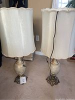 Pair of beige and gold glass lamps with scalloped beige fabric shades, metal ornate claw foot bases, standing on carpeted floor.