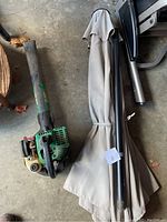 Photo of the folded beige market umbrella and gas-powered leaf blower side by side on a concrete floor.