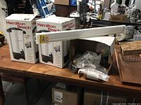 Two boxed Hamilton Beach coffee urns (42 cup), a fluorescent overhead light fixture, copper-plated tray, and baking accessories on a wooden table.
